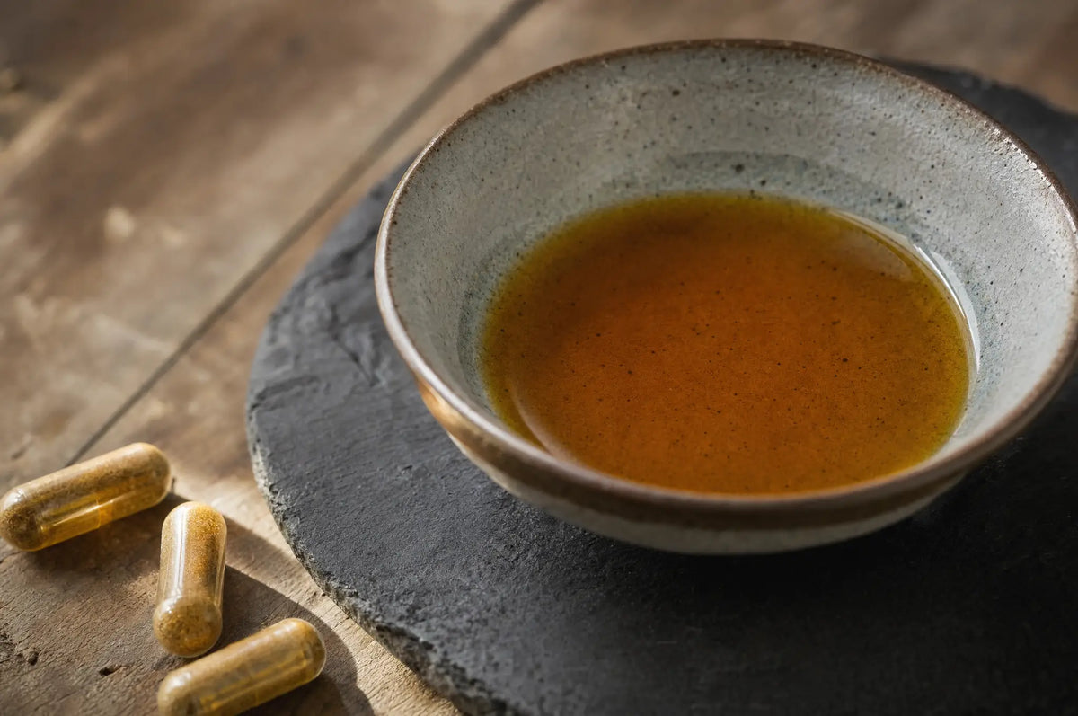 A small pour of amber apple cider vinegar pooling in a shallow glazed stoneware bowl on dark slate, beside loose capsules.