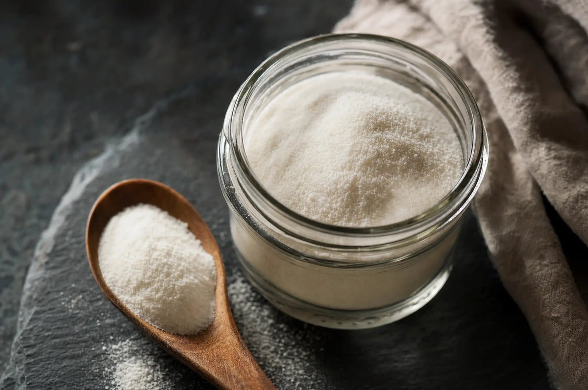 A small glass jar of unflavored collagen peptide powder on dark slate, with a wooden spoon beside it.