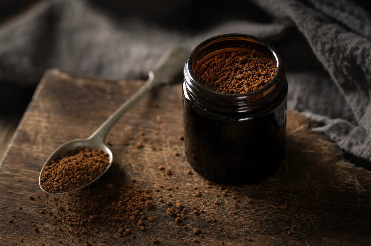 A single dark glass bottle of caffeine powder resting on a weathered wooden cutting board, with a metal spoon beside it.