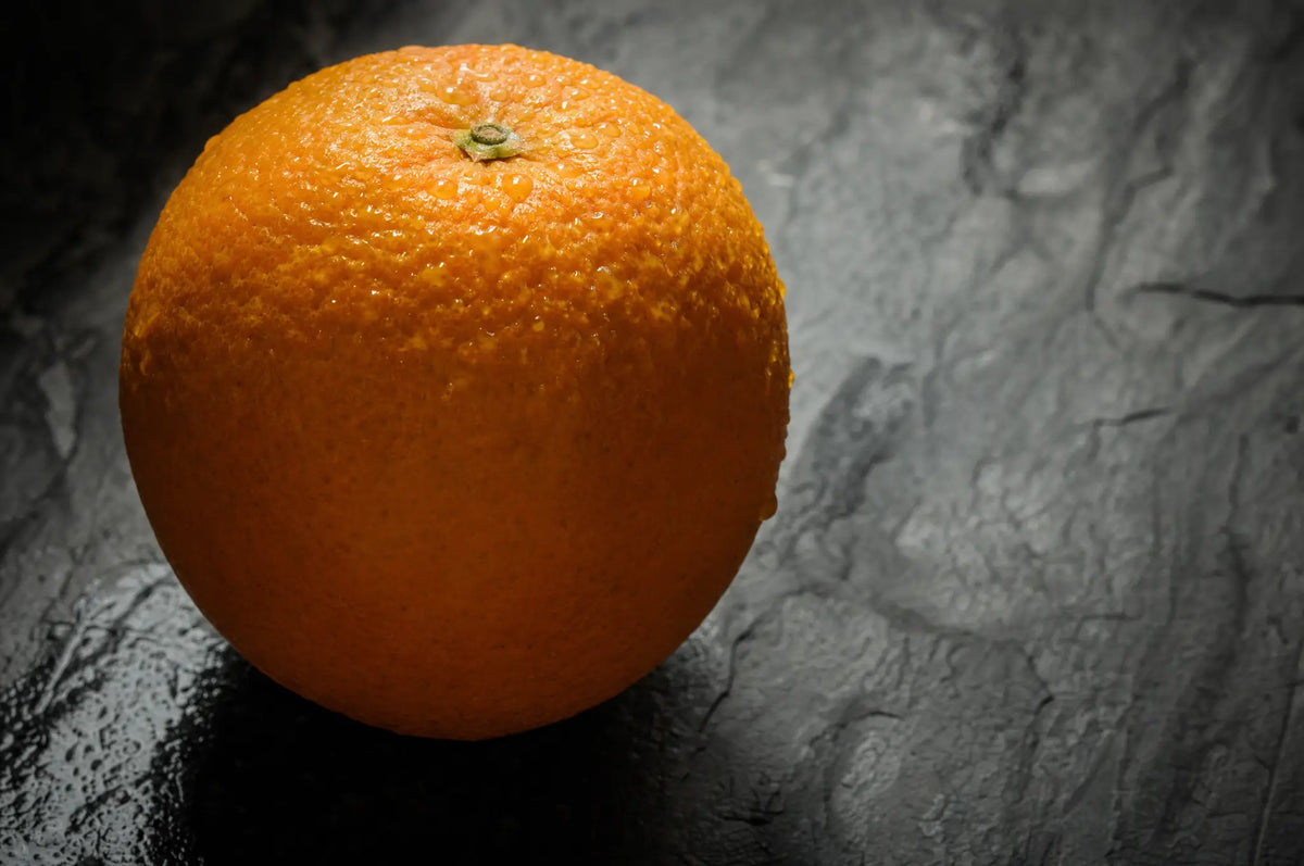 The “Catecholamine Honeymoon”: Why Strict Diets Feel Easy—Then Get Hard — A single raw orange on a dark slate surface, its peel dimpled and glistening with morning dew, side-lit to cas