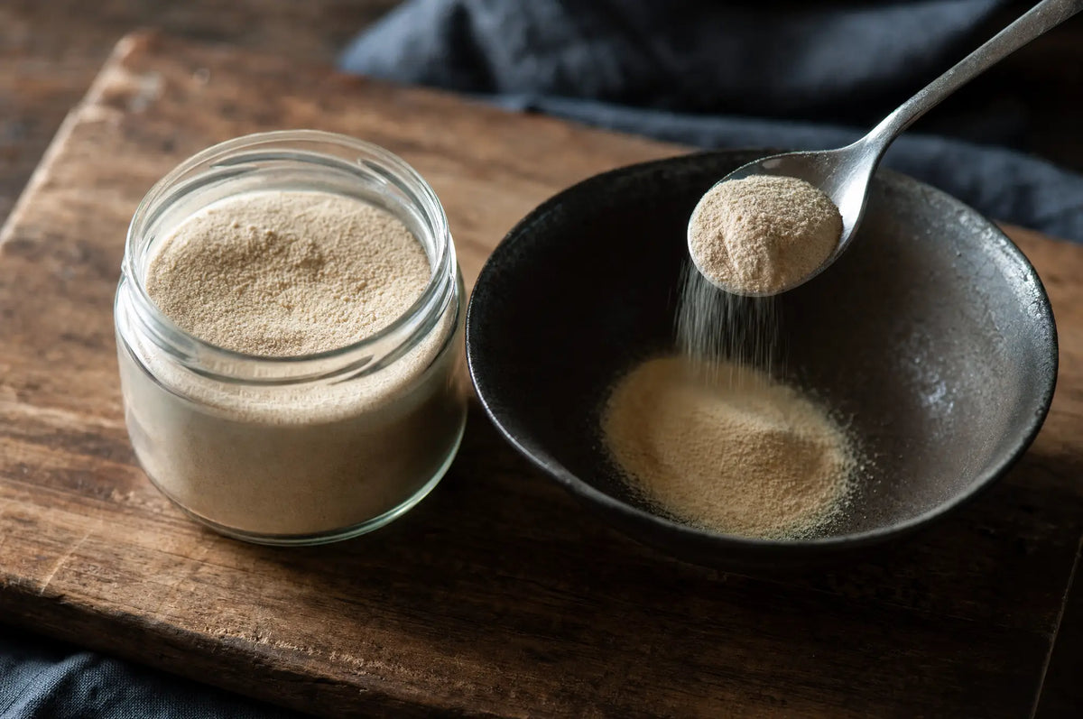 Small glass jar of beige collagen powder on a weathered cutting board, a metal teaspoon spooning powder into a ceramic bowl.
