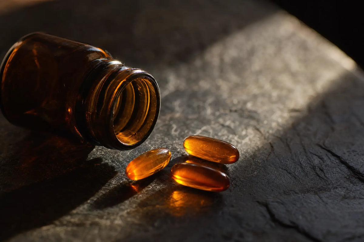 A small amber glass bottle tipped on its side on dark slate with three loose ubiquinone capsules beside it, soft window light