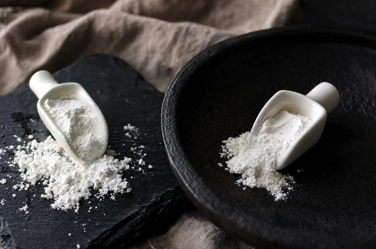 Two small white glycine powder scoops spilling onto dark slate beside a matte black cast iron surface, soft shadows.