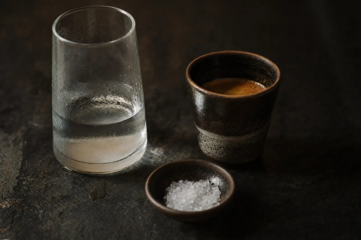 Hydration and Energy: Why Water Helps—But Cannot Explain Every Slump — A half-drunk glass of water on a dark slate surface beside a single espresso cup and a small bowl of loose sal
