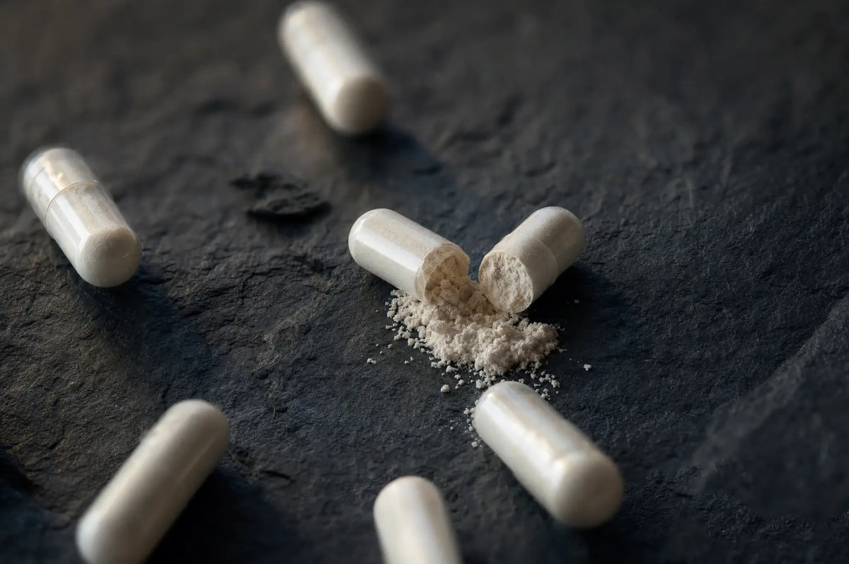 Loose magnesium glycinate capsules spilled on dark slate, one capsule split open to reveal powder inside.