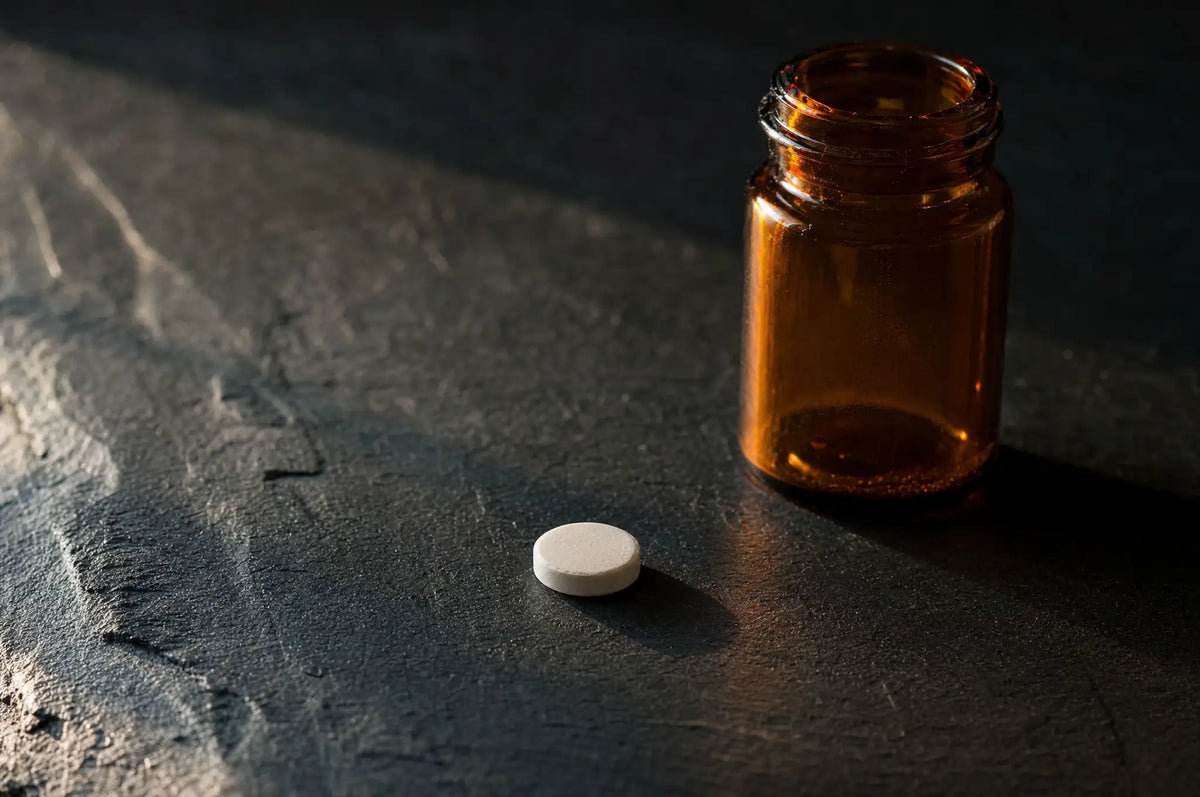 Melatonin Dose: Why More Is Not Always Better — A single white melatonin tablet resting on a dark slate surface beside a small amber glass bottle, soft side l