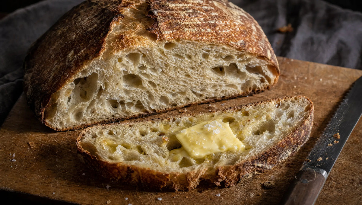 A halved sourdough loaf with an open crumb and a buttered slice on a wooden board