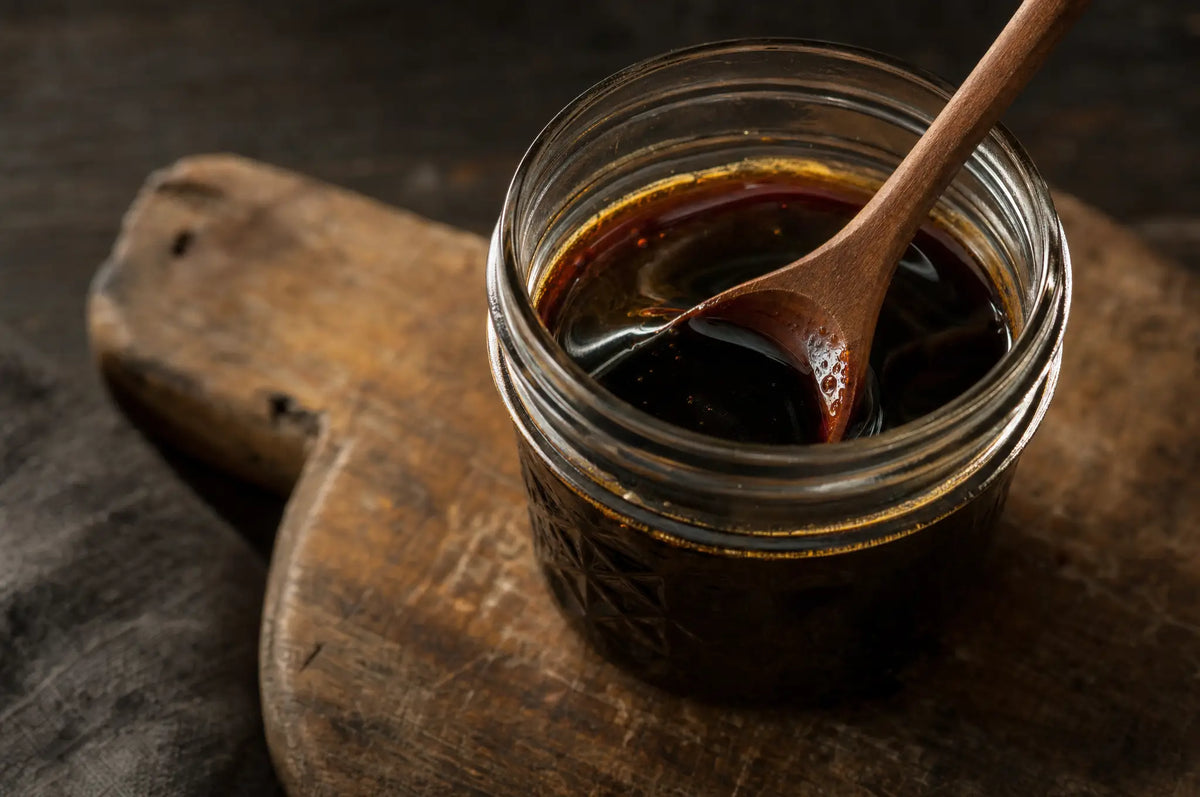 A glass jar of dark blackstrap molasses with a wooden spoon, on a worn wooden cutting board
