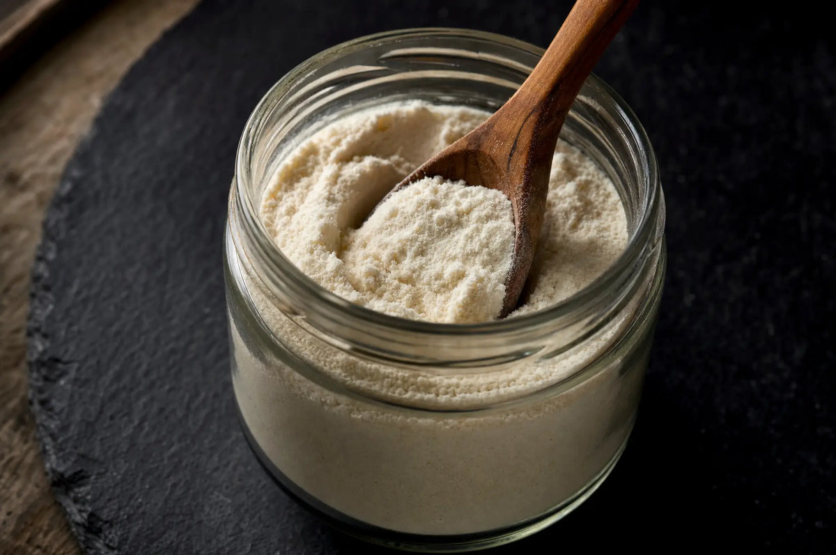 The Best Time of Day to Take Collagen for Absorption — A small glass jar of creamy collagen powder resting on dark slate, with a wooden spoon partially submerged, na