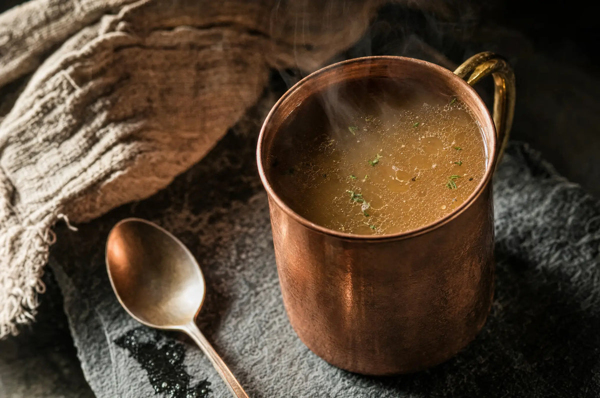A copper mug of steaming beef bone broth on dark slate with a spoon resting beside it, capturing metabolic recovery.